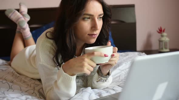 Young woman drinking coffee using computer lying on her bed alt