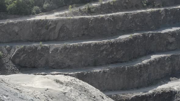 The high terraces of a dusty, gray quarry towering above one single excavator at the bottom. Handhel alt