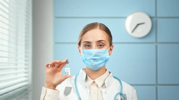 Young woman doctor in blue disposable face mask and white coat holds vial alt