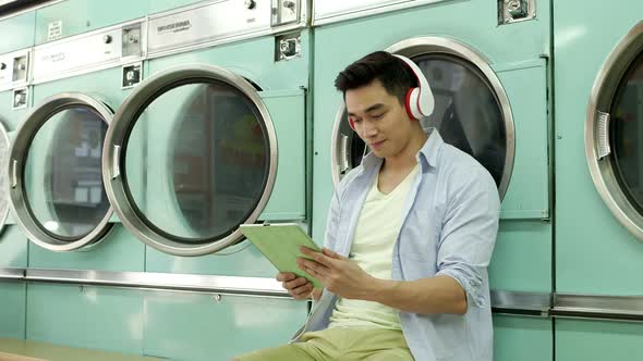 A Young Man sits using his Digital Tablet whilst waiting for his laundry