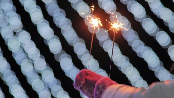Sparklers in the Hands of a Girl in Mittens Against the Background of the City's Christmas Lights alt
