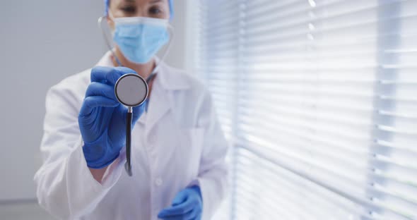 Caucasian female doctor wearing face mask and surgical gloves using stethoscope alt