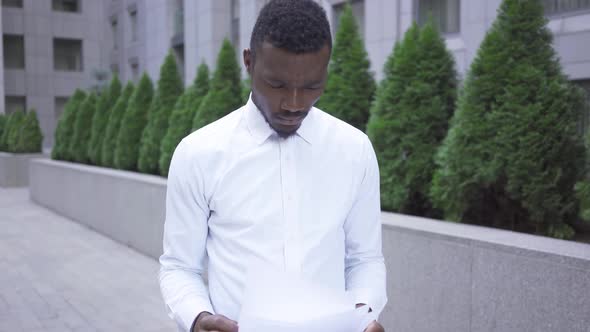 Troubled Young African American Man in White Shirt Looking Through Papers Standing on the City alt