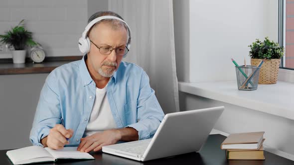 An Elderly Man in Headphones and a Computer Takes Notes in a Notebook