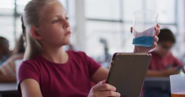 Caucasian schoolgirl using tablet looking at blue liquid in jar during chemistry class alt