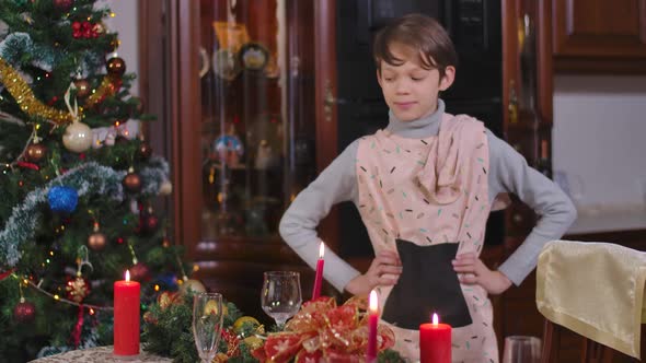 Satisfied Brunette Caucasian Boy Putting Hands on Hips Admiring Decorated Christmas Table alt