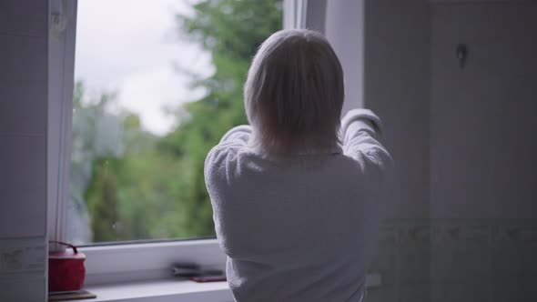 Back View Confident Caucasian Senior Woman with Grey Hair in White Bathrobe Stretching Looking Out alt