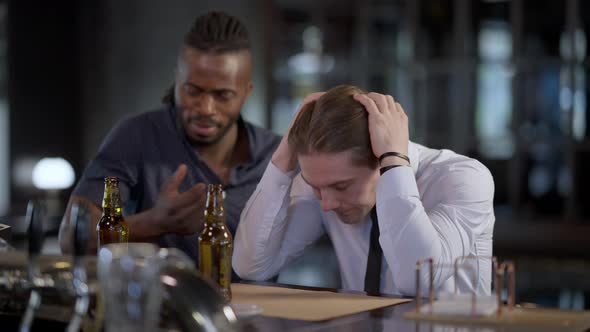 Frustrated Caucasian Man Holding Head in Hands As African American Friend Cheering Up Mate alt