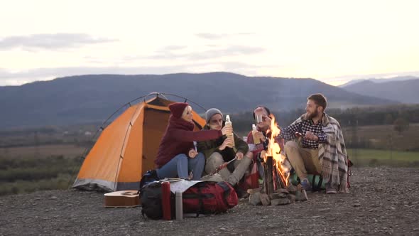 friends having rest near tourist tent with bonfire on which fried sausages in mountains alt