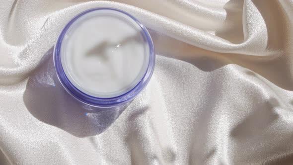 Blue glass cream jar on smooth silk background in sunlight with flowers shadow 4K skincare cosmetics alt
