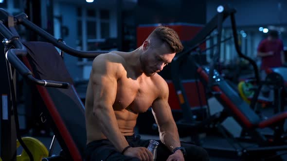 Handsome bodybuilder man drinks water in gym. Male athlete rests on bench. Slow motion. alt