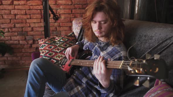 Portrait of Young Man Musician with Long Red Hair Playing Bass Guitar at Home Sitting on Sofa alt