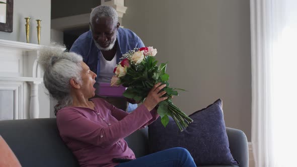 African american senior man giving gift box and flower bouquet to his wife at home alt