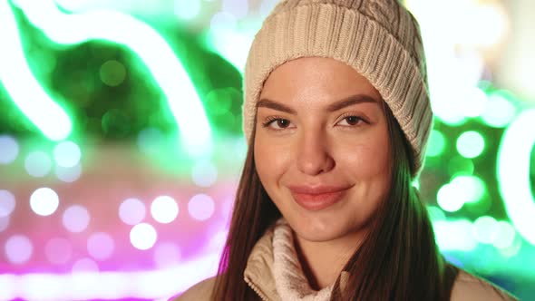 Portrait of Joyful Caucasian Woman in Hat Looking at Camera Smiling Around Christmas Lights alt