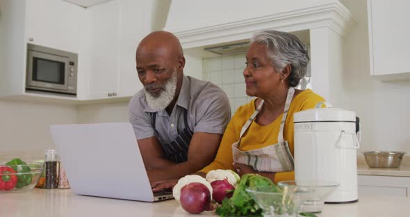 African american senior couple wearing aprons using laptop together in the kitchen at home alt