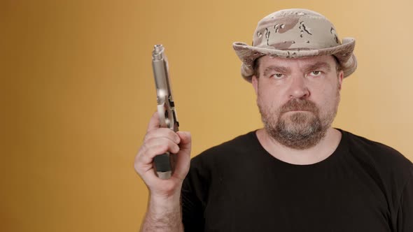 Middle aged man with gun in black t-shirt and hat against yellow background. alt