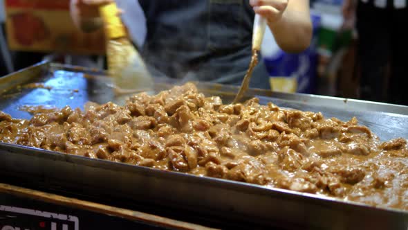 Chef stirring meat stew on an open flat grill, train night market Bangkok alt