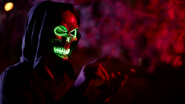 Portrait of a Person in a Glowing Mask of Death and in a Hood Against a Background of Branches alt