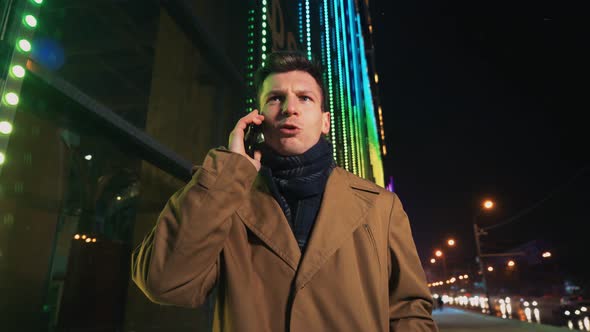 Angry Man Talking on Mobile Phone Outside Glowing Shopping Mall at Night alt