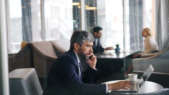 Entrepreneur in Talking on Mobile Phone and Using Laptop Working in Cafe alt
