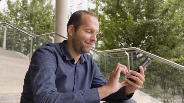 A Caucasian Man Works on a Smartphone with a Smile As He Sits on a Staircase in an Urban Area