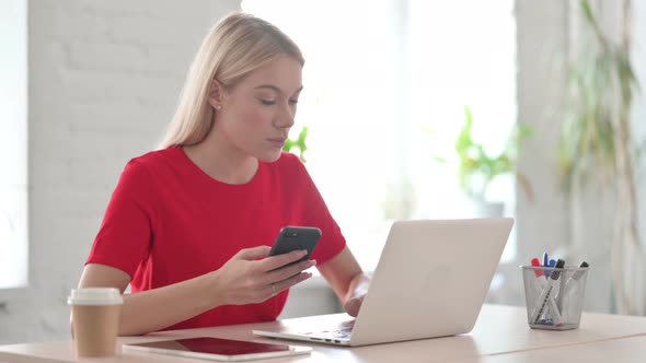 Young Blonde Woman Browsing Internet on Smartphone while using Laptop in Office alt