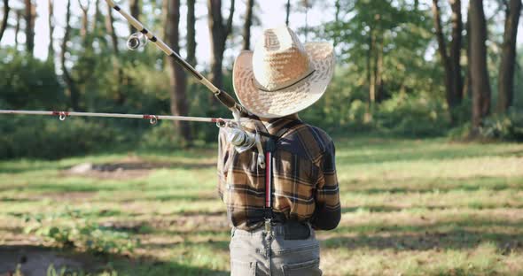 Boy in Straw Hat which Holding Fishing Rods on His Shoulders when Goes Fishing Among Green Trees alt