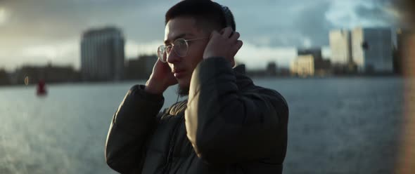 A guy listening music holding the headphones with his hands next to the river and slowly dancing alt