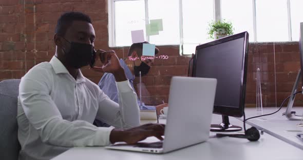 African american businessman wearing mask sitting on desk using laptop talking on smartphone alt