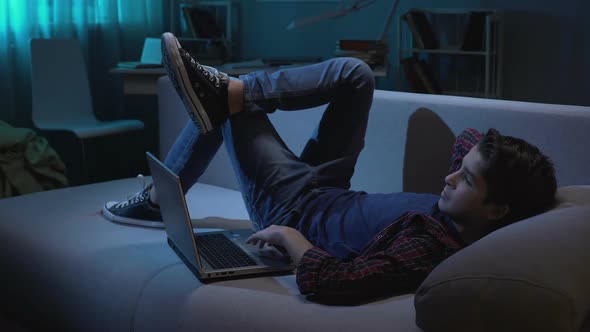 Teenage Boy Lying on Sofa, Looking Around and Watching Adult Videos on Laptop alt