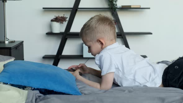 Cute cheerful little child playing video games on phone lying on a big white bed