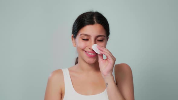 A Young Brunette Girl Rubs Her Face with a Cotton Pad on a White Background