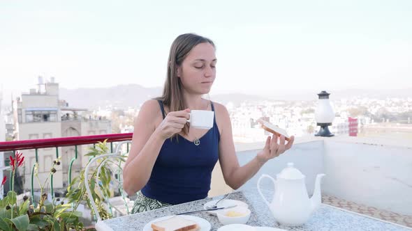 Woman is Having a Breakfast in Cafe on Terrace on City and Mountain Background
