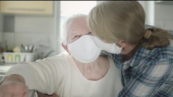 Old Woman Hugging Her Daughter In The Kitchen. Both Women In Medical Masks To Protect alt