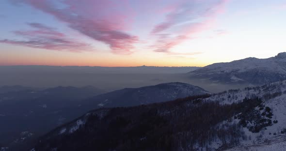 Backward Aerial Top View Over Winter Snowy Mountain and Woods Forest at Sunset or Sunrise