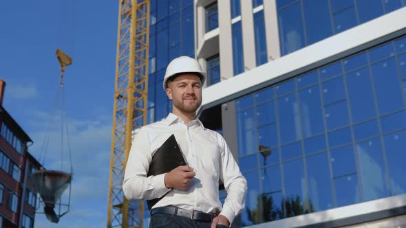 Architect Engineer in a White Shirt and Helmet on a Background of a Modern Glass Building Holds a alt
