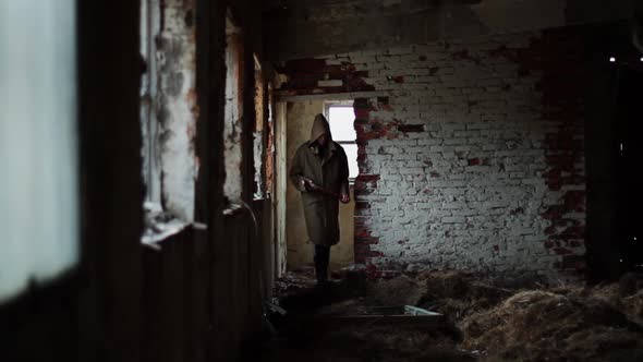 Silhouette of Man in Dark Cloak Walking Through Abandoned Building with an Ax alt