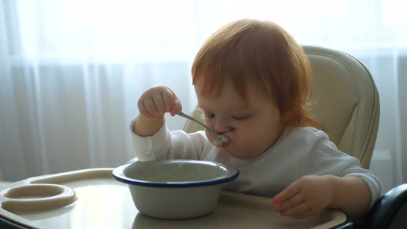 Close-up of a Baby Sitting at the Table and Eating Baby Food with a Spoon on Its Own