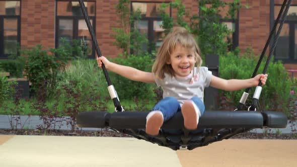Little Girl Swing on Swing Looks at Camera Laughs and Smiles on Background on Playground alt
