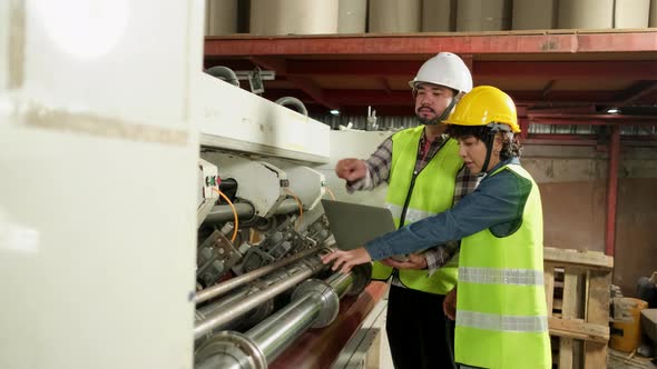 Two industrial workers use a laptop to control a paper manufacturing machine. alt