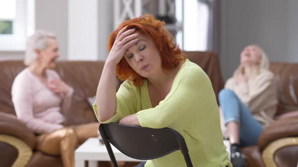 Displeased Redhead Woman Sitting on Chair Shaking Head Talking As Blurred Cheerful Friends Laughing alt