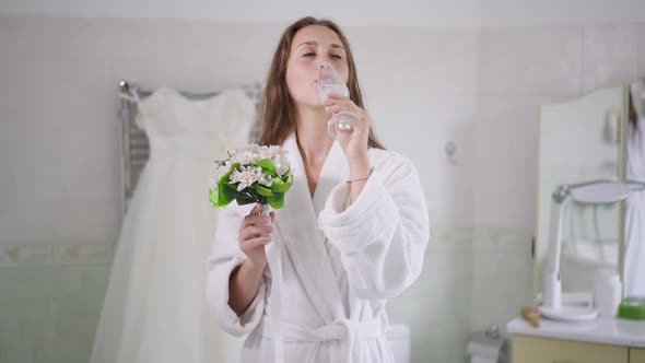 Front View of Smiling Charming Young Caucasian Bride Drinking Champagne Smelling Bouquet of Flowers alt