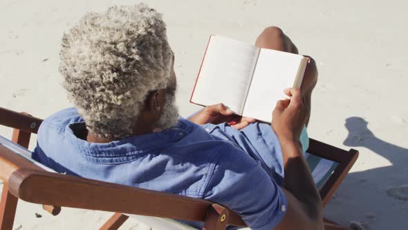 Senior african american man reading and lying on sunbed on sunny beach alt