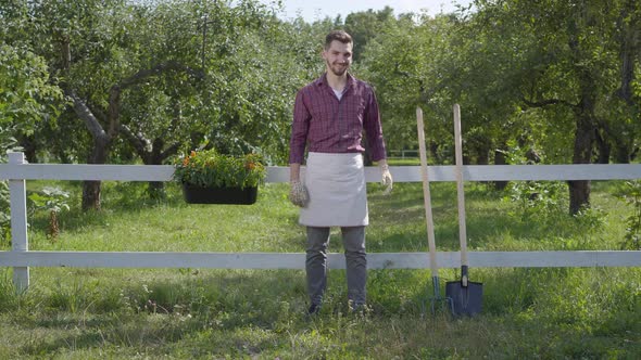 Professional Young Farmer in Garden Gloves Standing in the Green Summer Garden Near the Fence alt