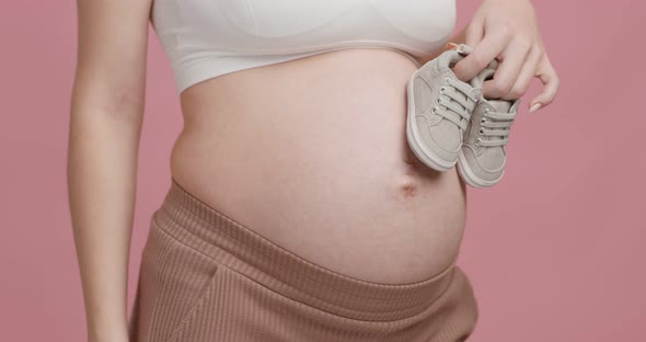 Close Up Shot of Pregnant Woman Holding Tiny Boots Near Her Big Belly Pink Studio Background alt