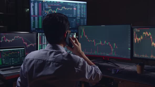 Back View Of A Asian Young Male Stock Market Broker Talking On Mobile Phone With Analyzing Graphs alt