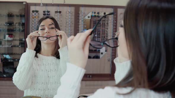 Young Brunette with Blue Eyes in a White Cozy Sweater Wears Glasses in Front of a Mirror in an alt