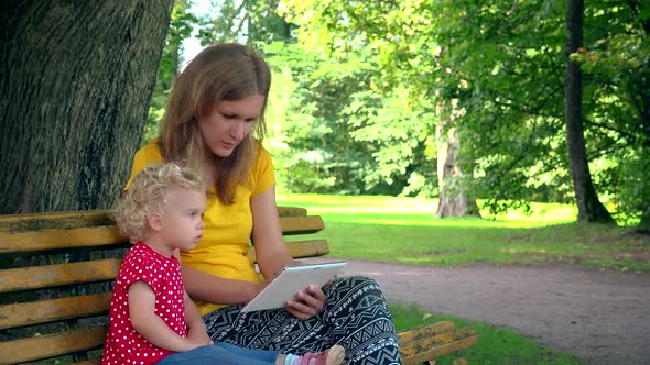 Mother With Toddler Daughter Girl Using Tablet Computer Sit On Bench In Park