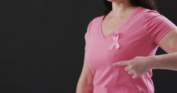 Mid section of a woman pointing to the pink ribbon on her chest against black background alt