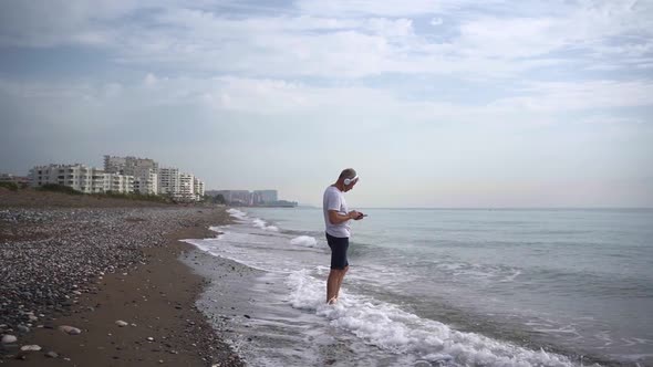 A Slender Whitehaired Elderly Man with Headphones Listens to Music on His Phone While Standing